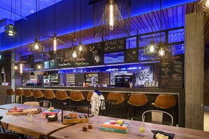 A modern bar area with wooden tables and chairs, a menu board, and hanging pendant lights. - Moxy Dresden Neustadt (Dresden)