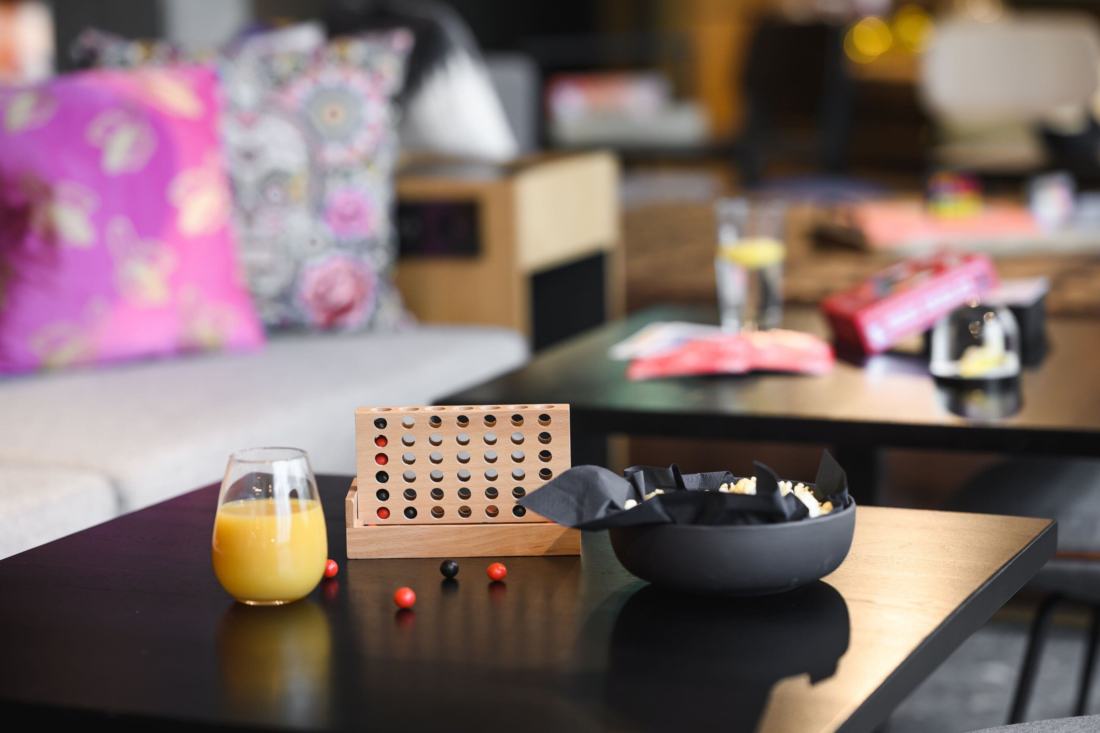 A modern living room with a coffee table, a glass of orange juice, a wooden puzzle board, and a bowl of snacks.