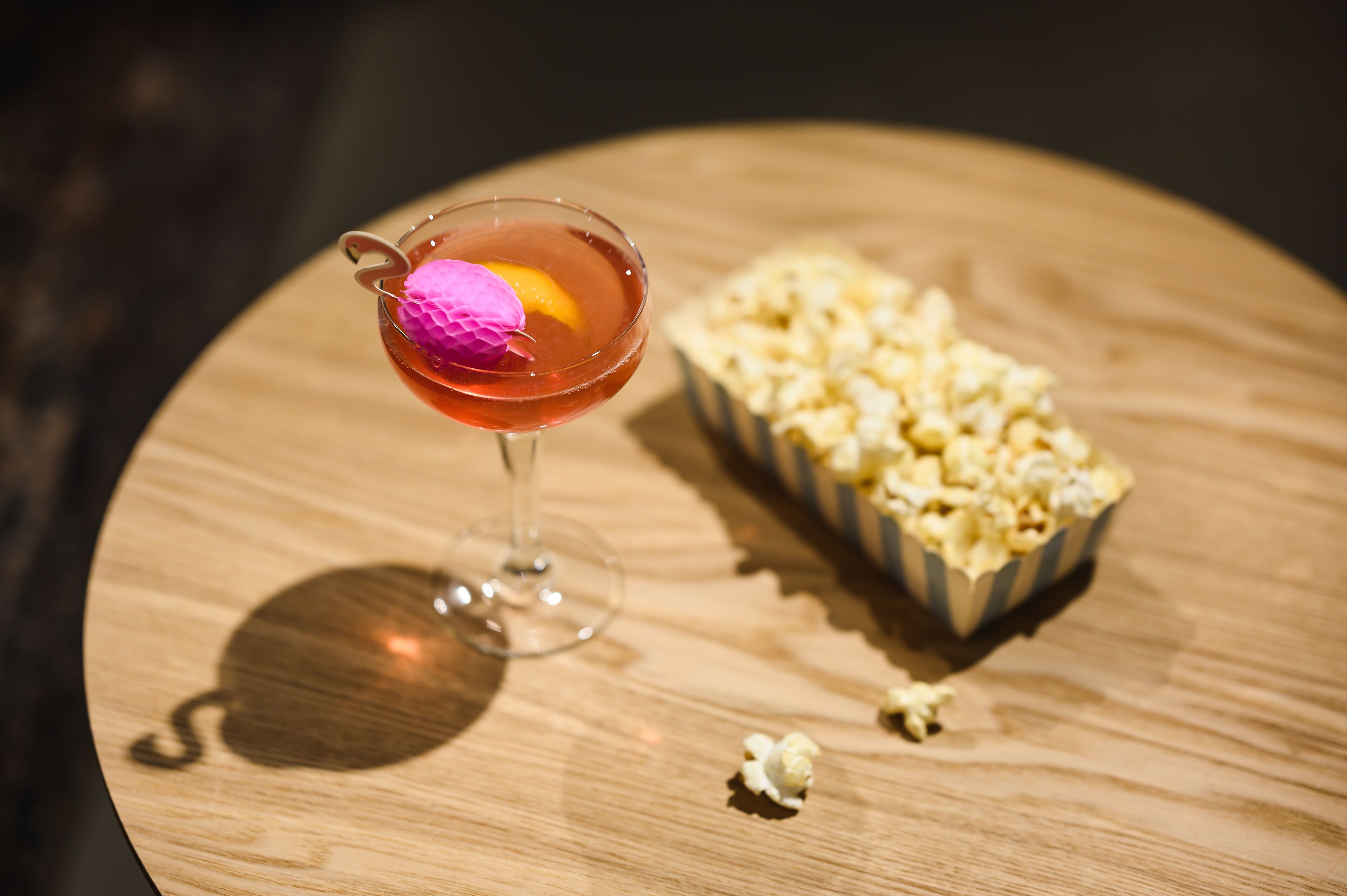 Una copa con un cubo de hielo rosa y una rodaja de naranja, además de una caja de palomitas sobre una superficie de madera.
