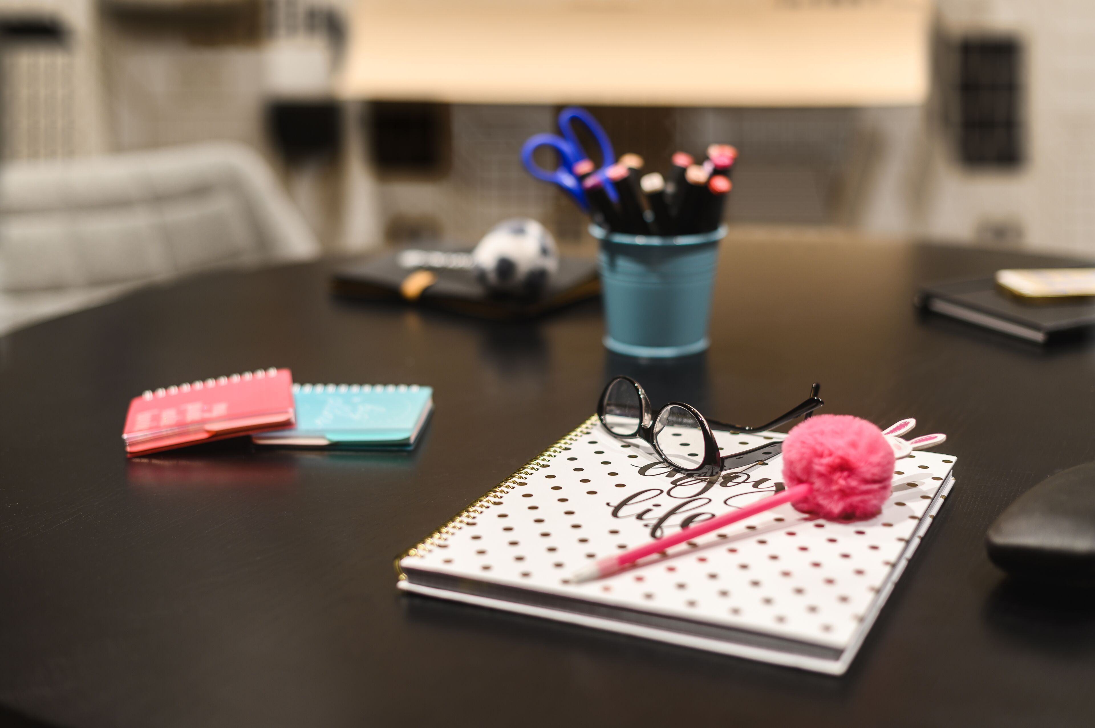 A round table with a notebook, a pen, a pair of glasses, a pink pom-pom, and a cup with pens and scissors.