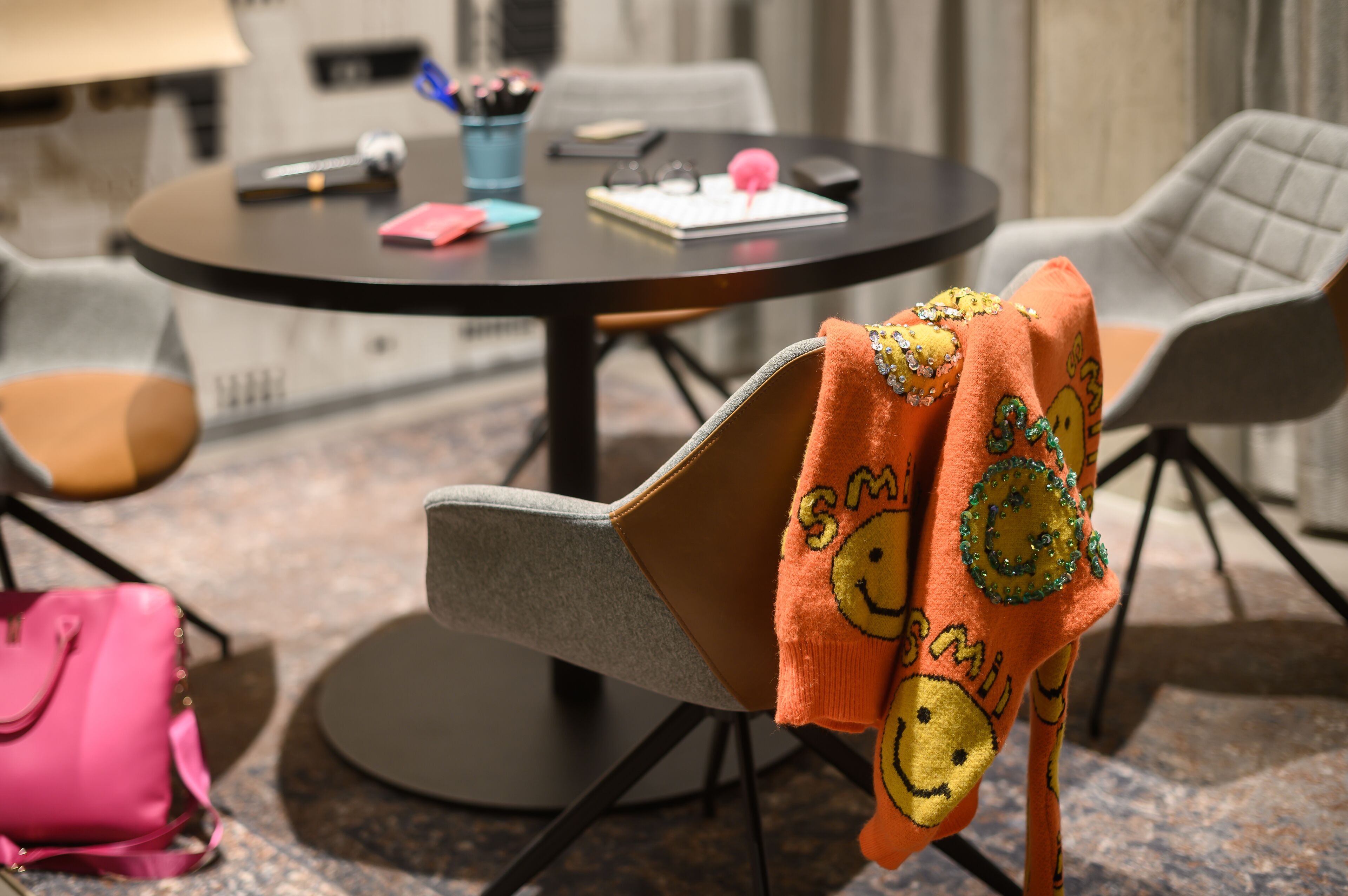 Une table ronde avec un carnet, des stylos et un objet rose. Une chaise est placée à côté, recouverte d’une couverture ornée de motifs représentant des visages souriants. Un sac rose est posé sur le sol.