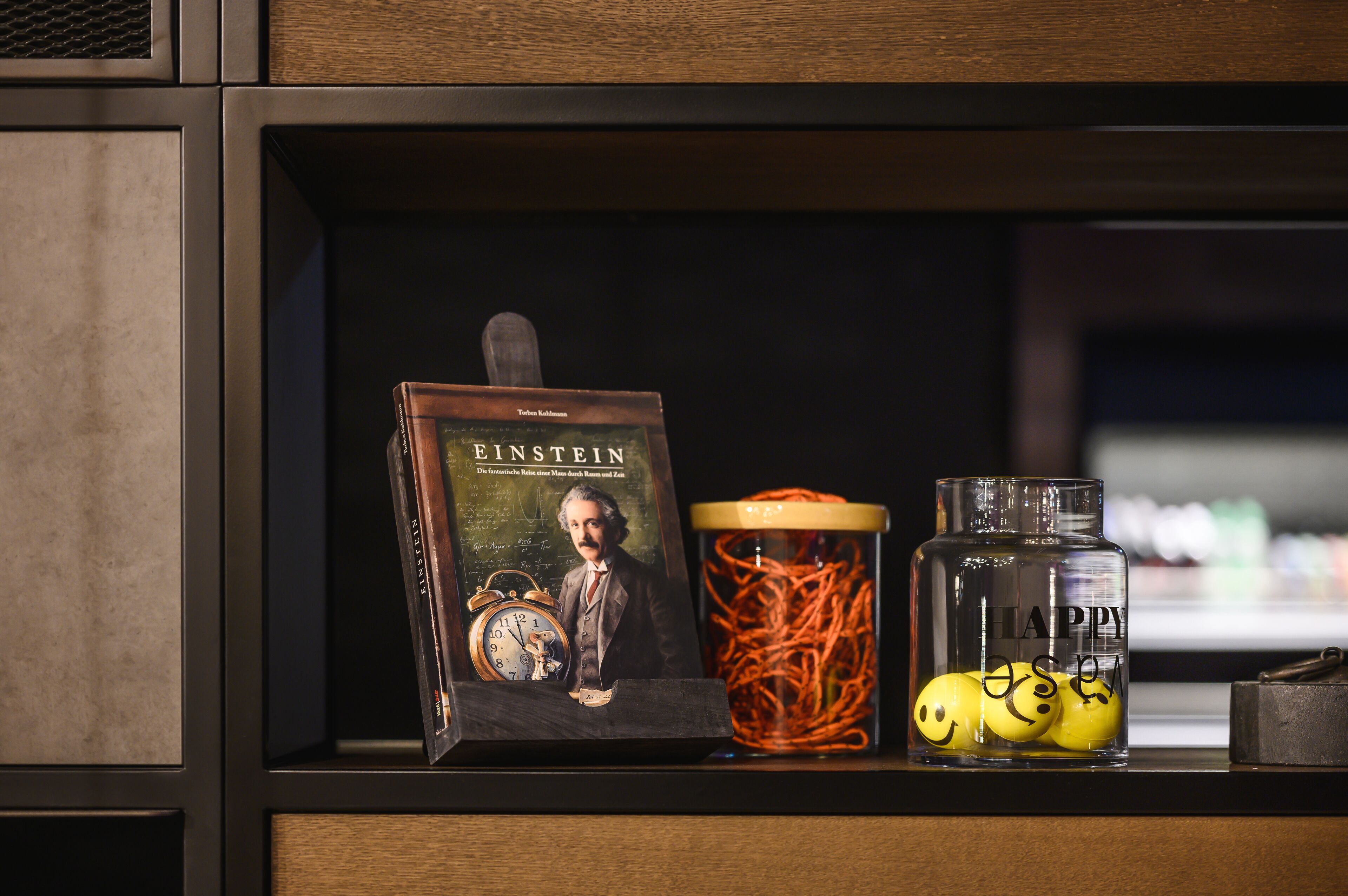 A shelf with a book titled 'Einstein', a glass jar with tangled orange strings, and a glass container with yellow balls.