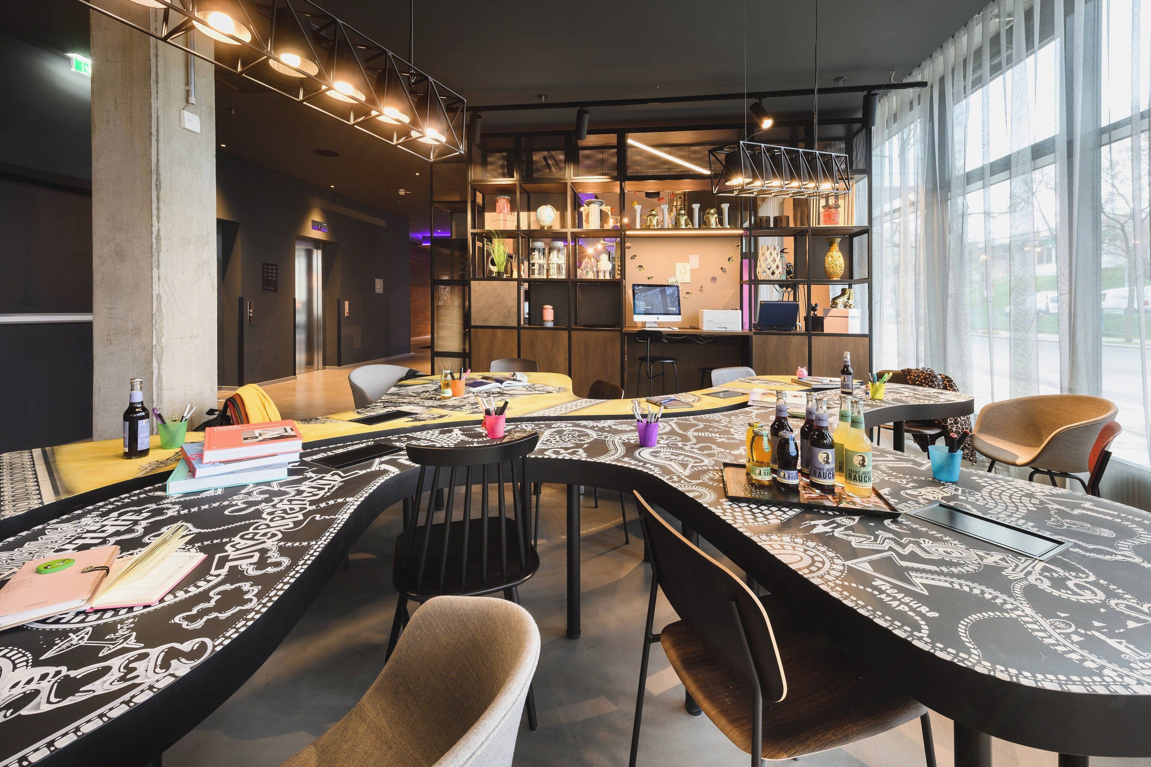 Uma sala de jantar moderna com uma mesa comunitária grande, diversas cadeiras e diferentes opções de assento, incluindo um sofá. Estantes com objetos decorativos e um monitor de computador são visíveis ao fundo.