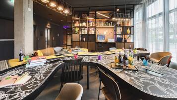 Un espace repas moderne avec une longue table conviviale, de nombreuses chaises et différents types de sièges, dont un canapé. On aperçoit des étagères ornées d’objets décoratifs et un écran d’ordinateur à l’arrière-plan.