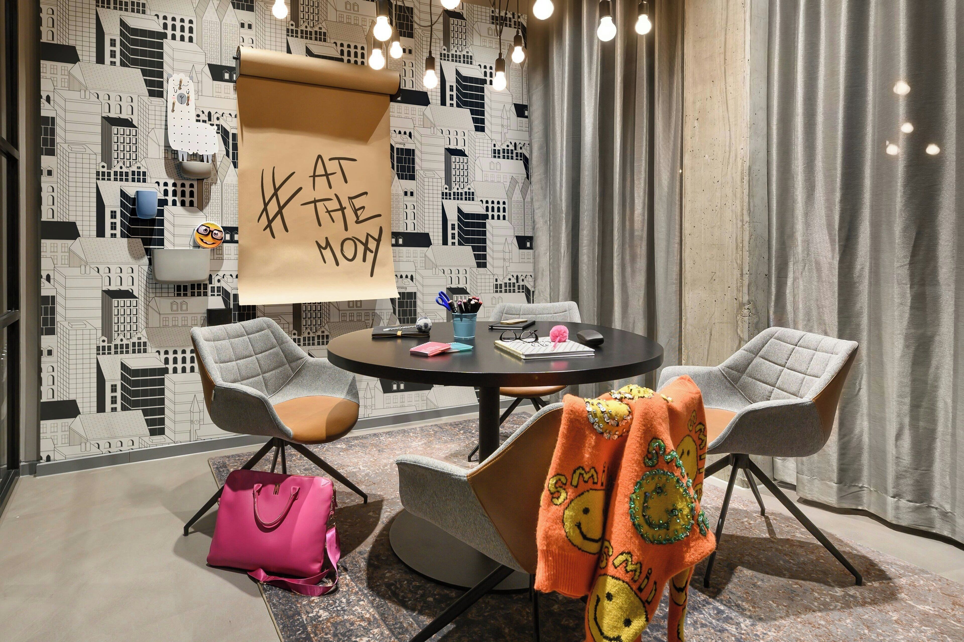 A modern room with a round table, two grey chairs, a pink handbag, and a colorful blanket on a chair. A banner with text hangs from the ceiling.