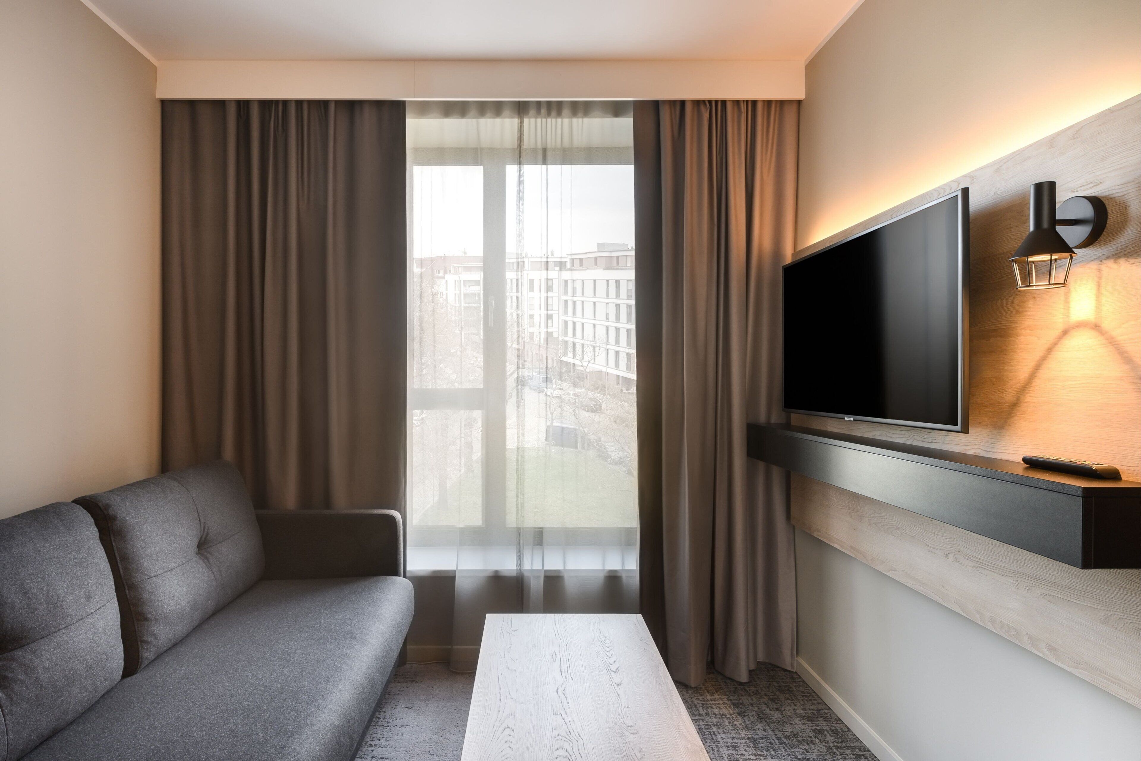 Ein modernes Hotelzimmer mit grauem Sofa, weißem Couchtisch, flachbildfernseher an der Wand und einem großen Fenster mit Vorhängen.