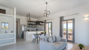 Un soggiorno moderno con un divano grigio, un tavolo da pranzo e sedie. C'è un angolo cottura con frigorifero, scaffali e una lampada a sospensione. Una porta scorrevole in vetro dà accesso a un patio esterno.