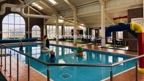 An indoor swimming pool with clear water, a diving board, and a caution sign on the right. - Above & Beyond Caravans4hire Lyons Robin Hood Rhyl (Rhyl)