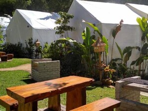 A garden area with wooden tables and benches, surrounded by white tents and tropical plants. - Maribaya Glamping Tent (Lembang)