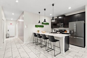 A modern kitchen with a white countertop, black bar stools, stainless steel appliances, and a backsplash with greenery.