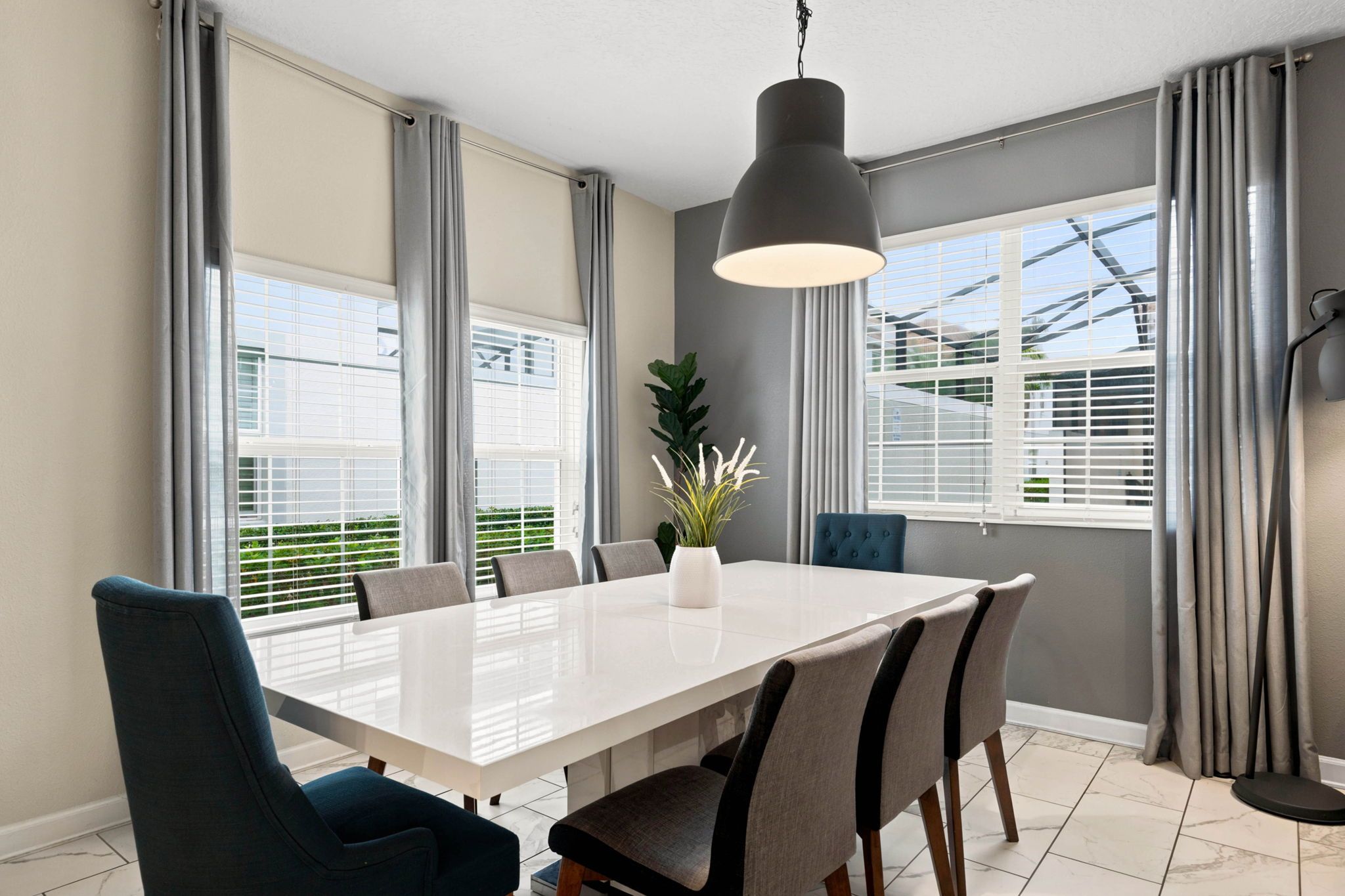 Une salle à manger moderne avec une grande table, des chaises, un luminaire suspendu et de grandes fenêtres équipées de stores.