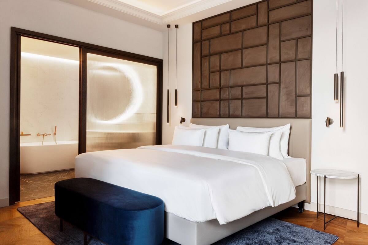 A modern bedroom with a large bed, a dark blue bench, a small round table, and a large window with a view of a cityscape.