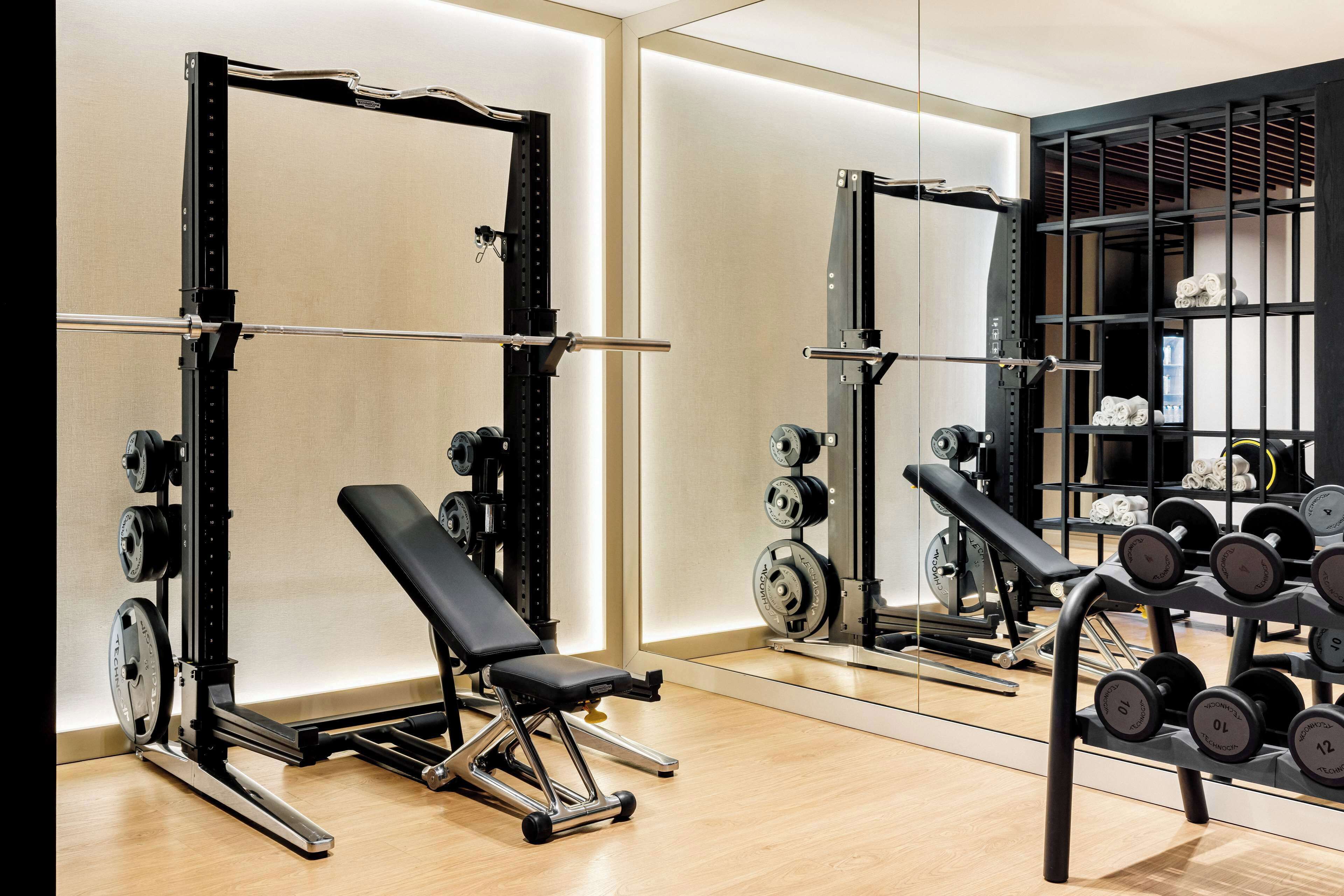 A gym with weightlifting equipment, a bench, and a mirror reflecting the equipment.