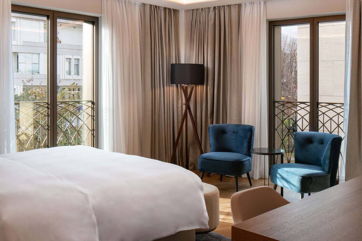 A hotel room with a large bed, two blue armchairs, a small table, and a floor lamp.