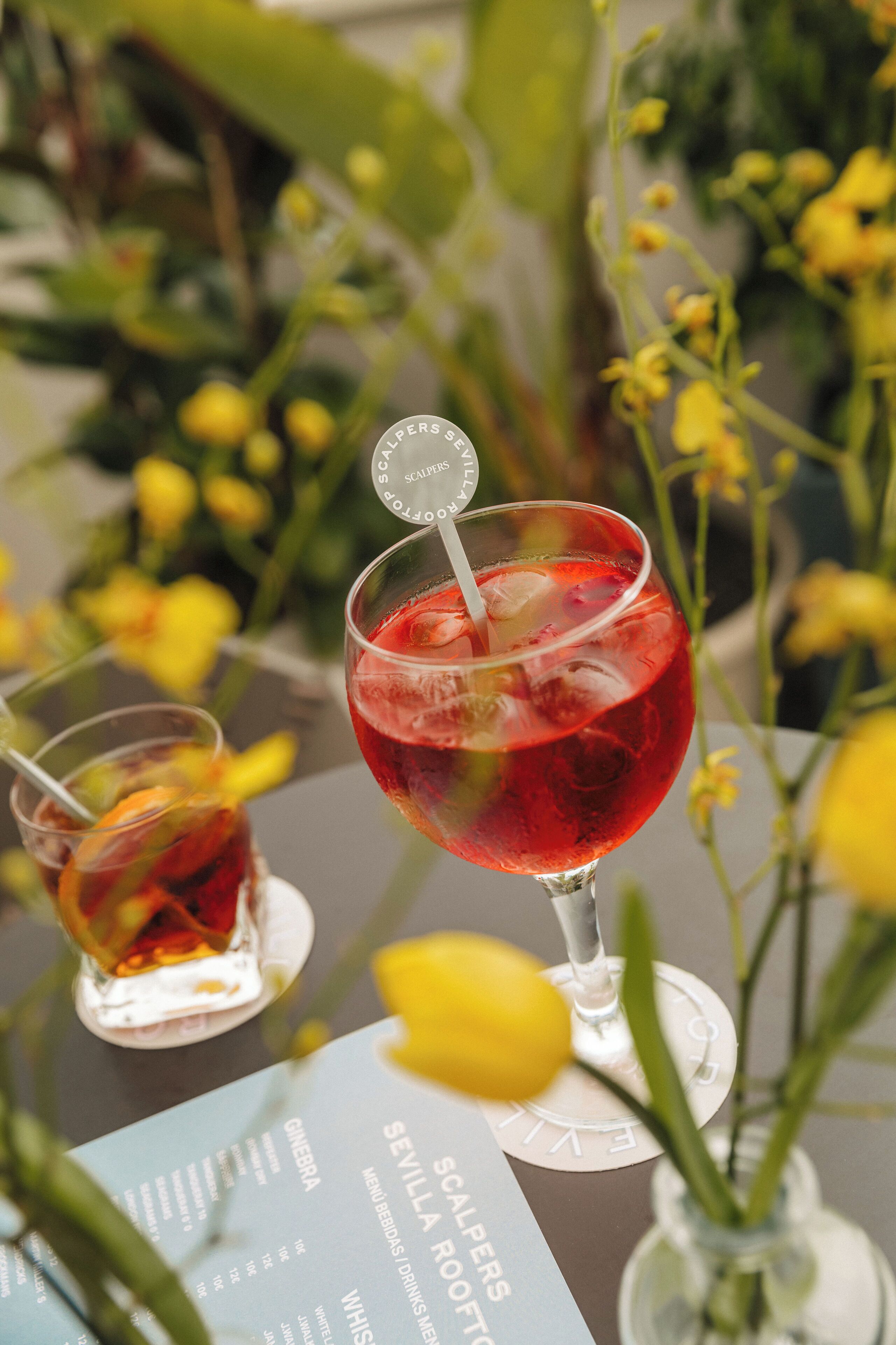 A table with two glasses of red beverage, a menu, and a vase with yellow flowers.