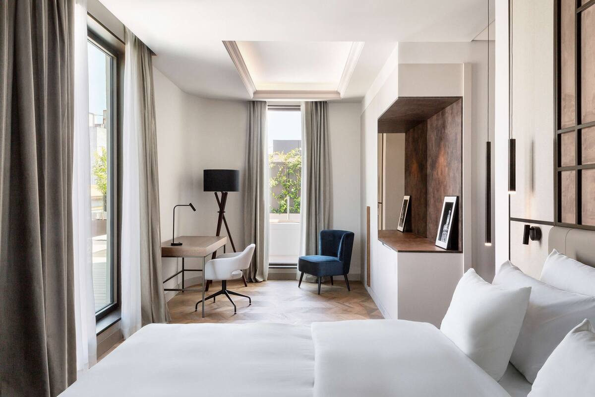A modern hotel room with a large bed, a desk, a chair, and a view of the outdoors.