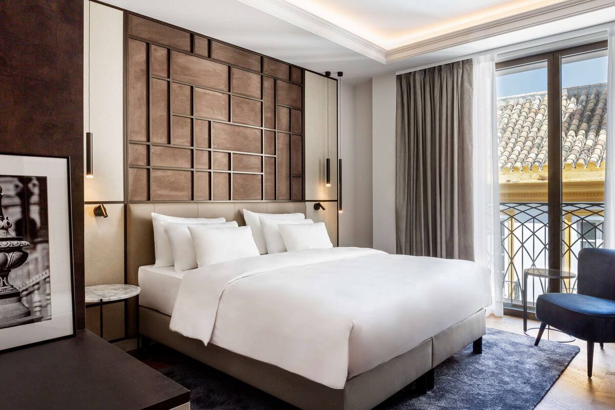 A modern hotel room with a large bed, a small side table, a framed picture, a dark-colored armchair, and a balcony with a view of the city.