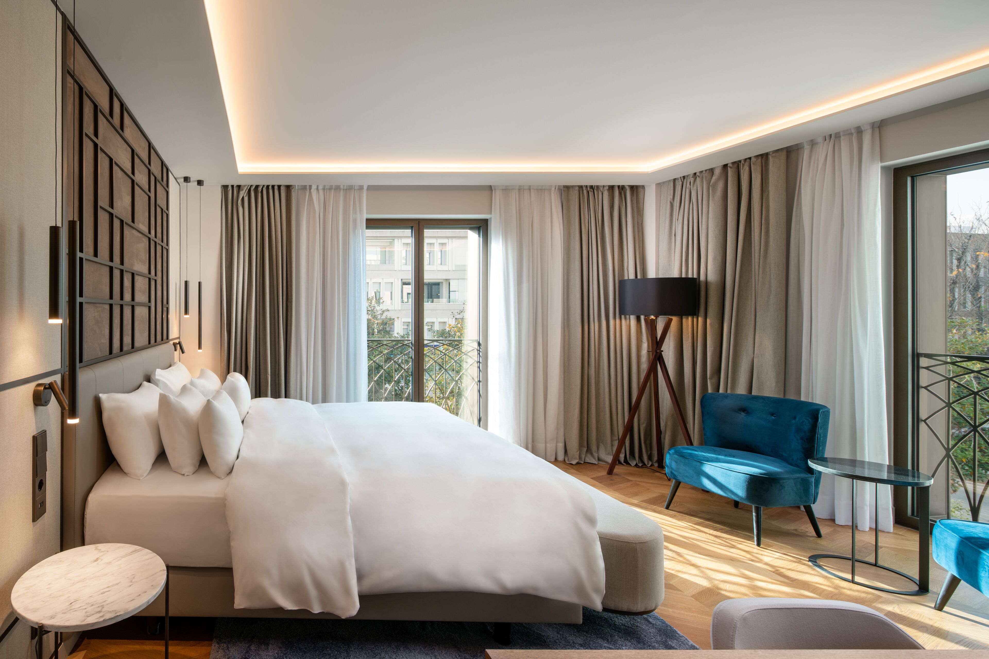 Ett modernt hotellrum med en stor säng, en sittgrupp med en blå fåtölj, ett litet runt bord och utsikt över staden genom stora fönster.