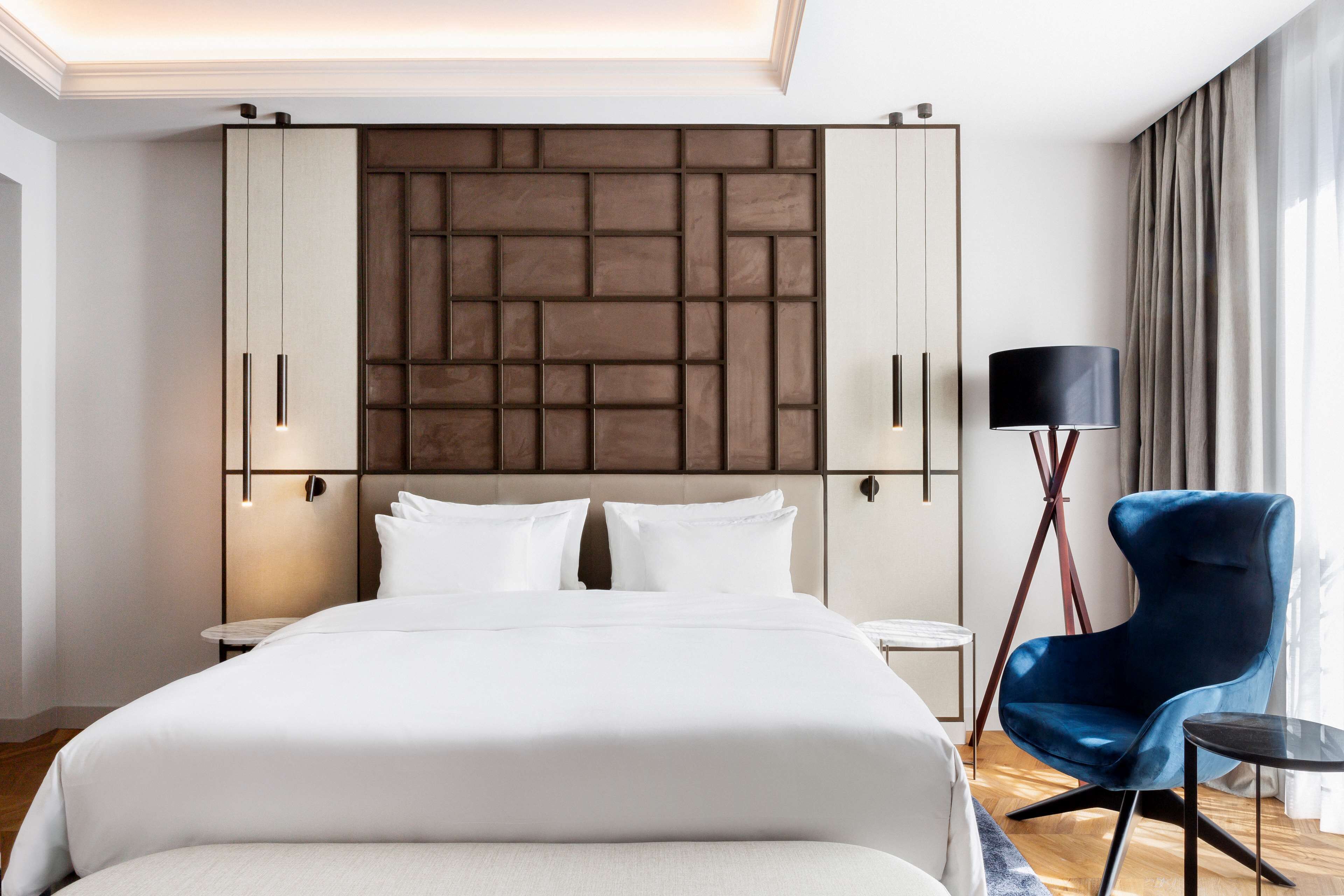 Una habitació d'hotel moderna amb un llit gran, un panell de capçal amb un disseny geomètric, un butaca blava, una taula petita i una làmpada.