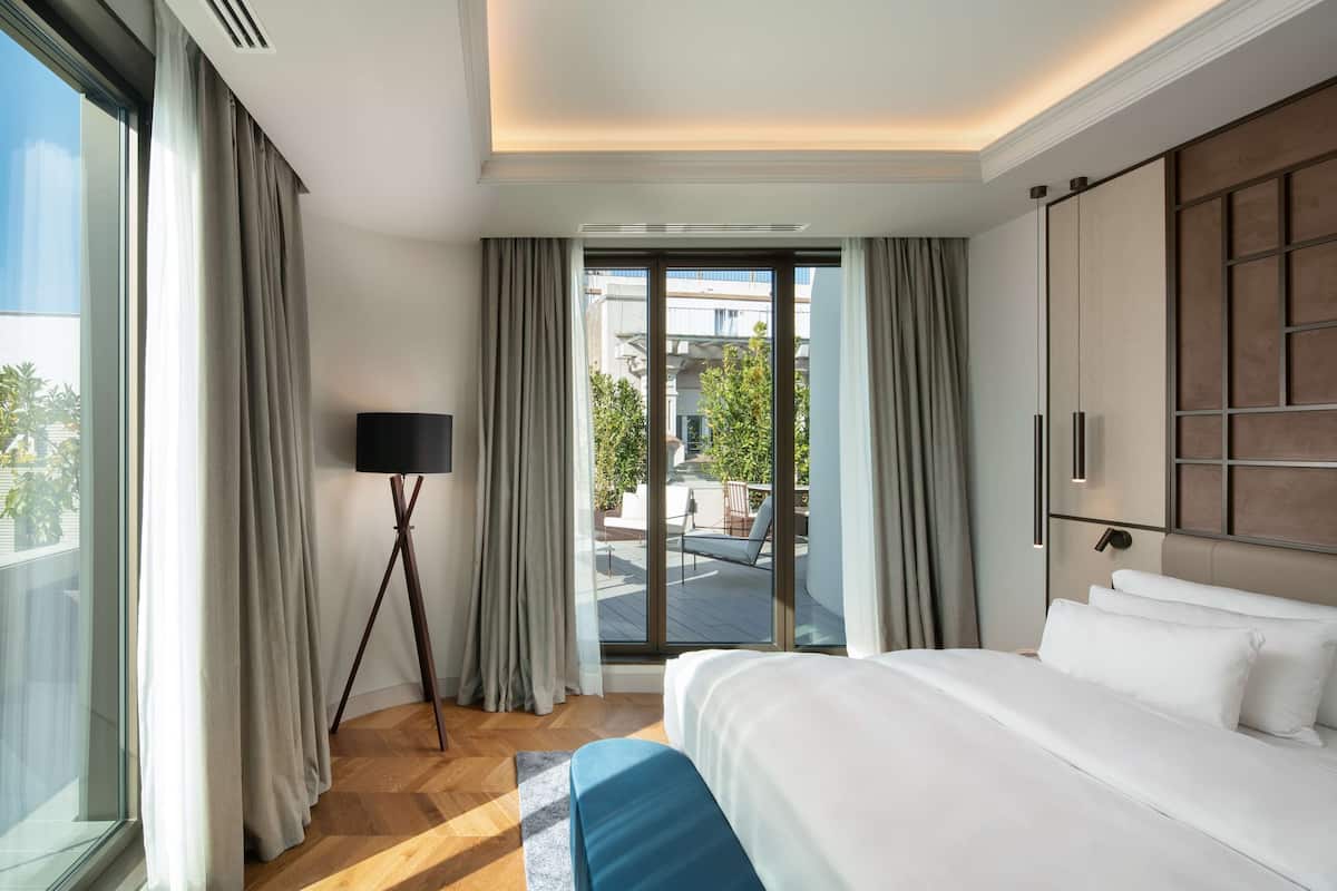 A modern hotel room with a large bed, a view of the outdoor balcony, and a floor lamp.