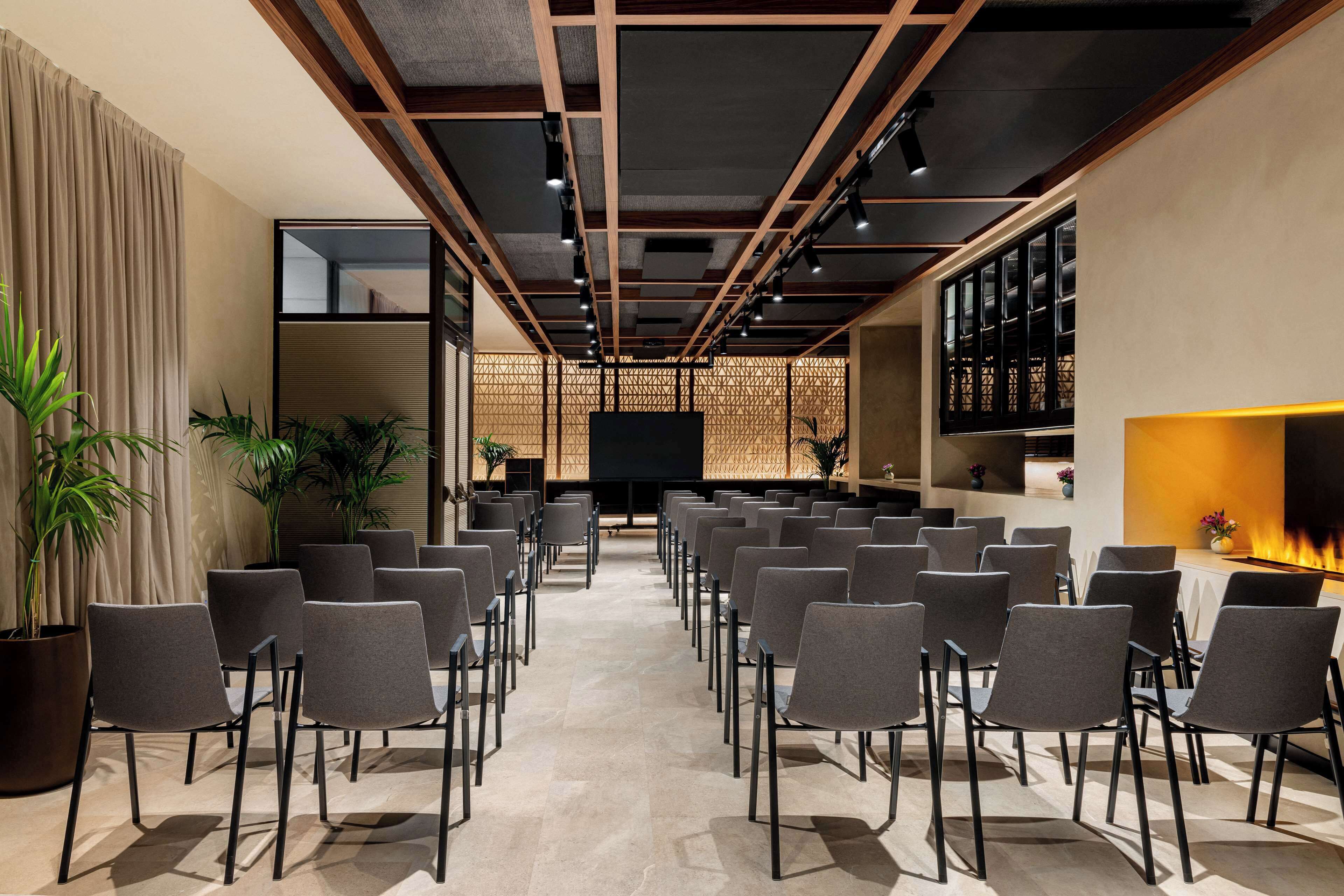 A conference room with rows of chairs facing a large screen, wooden beams on the ceiling, and a decorative wall with a fire feature.