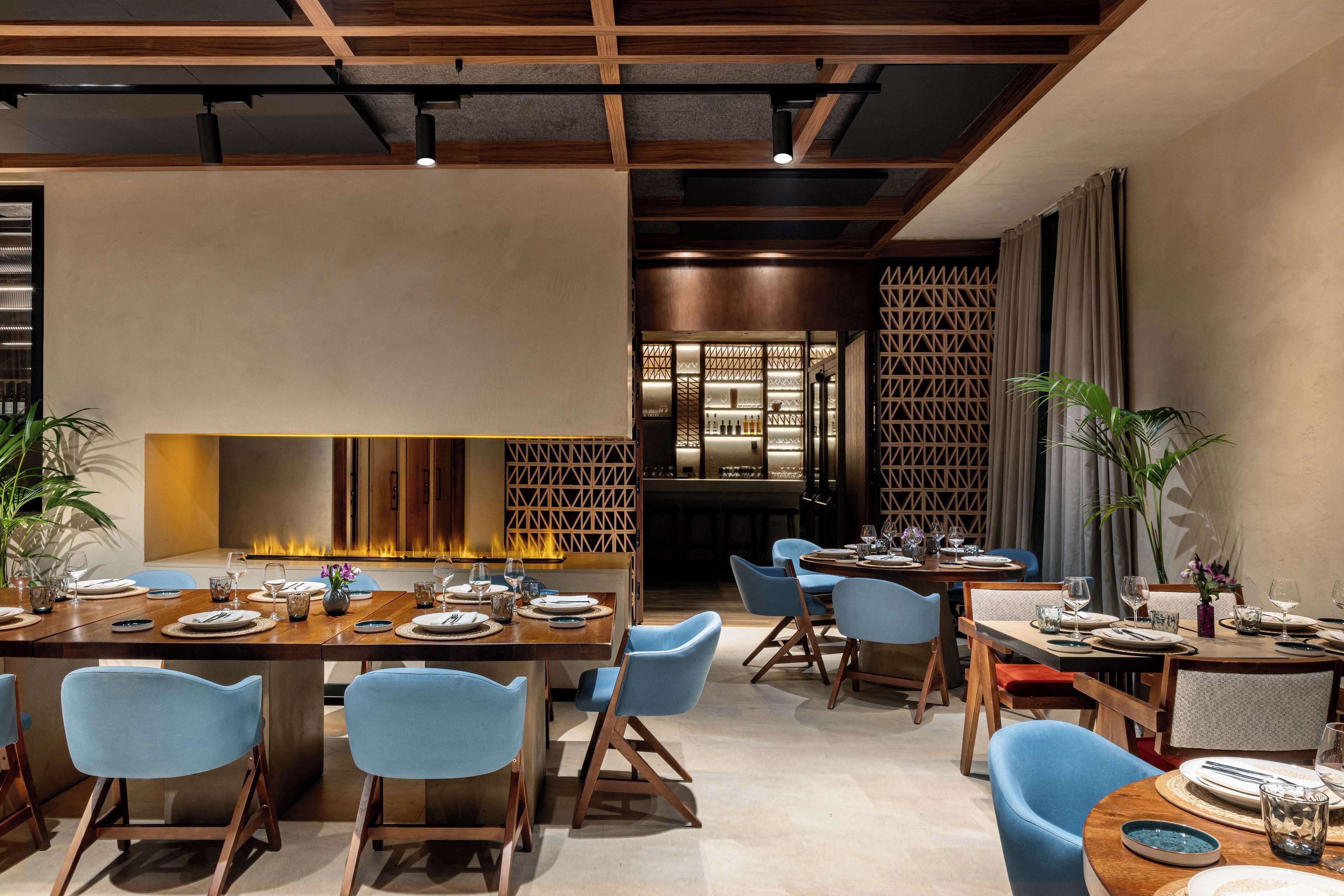 Un comedor moderno con mesas de madera y sillas azules, una chimenea central y una pared decorativa con patrones geométricos.