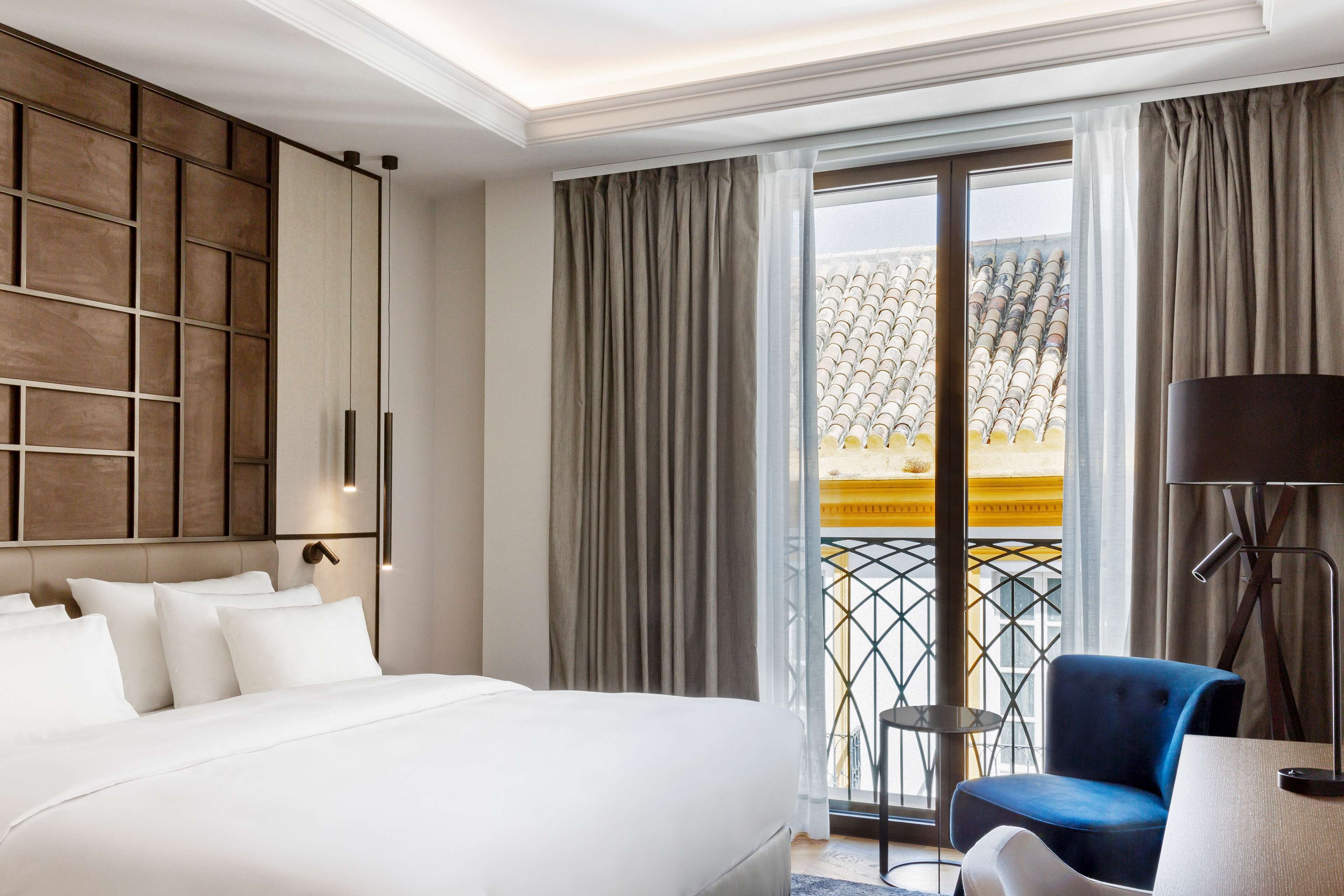 A hotel room with a large bed, a desk with a lamp, a blue chair, and a balcony with a yellow railing.