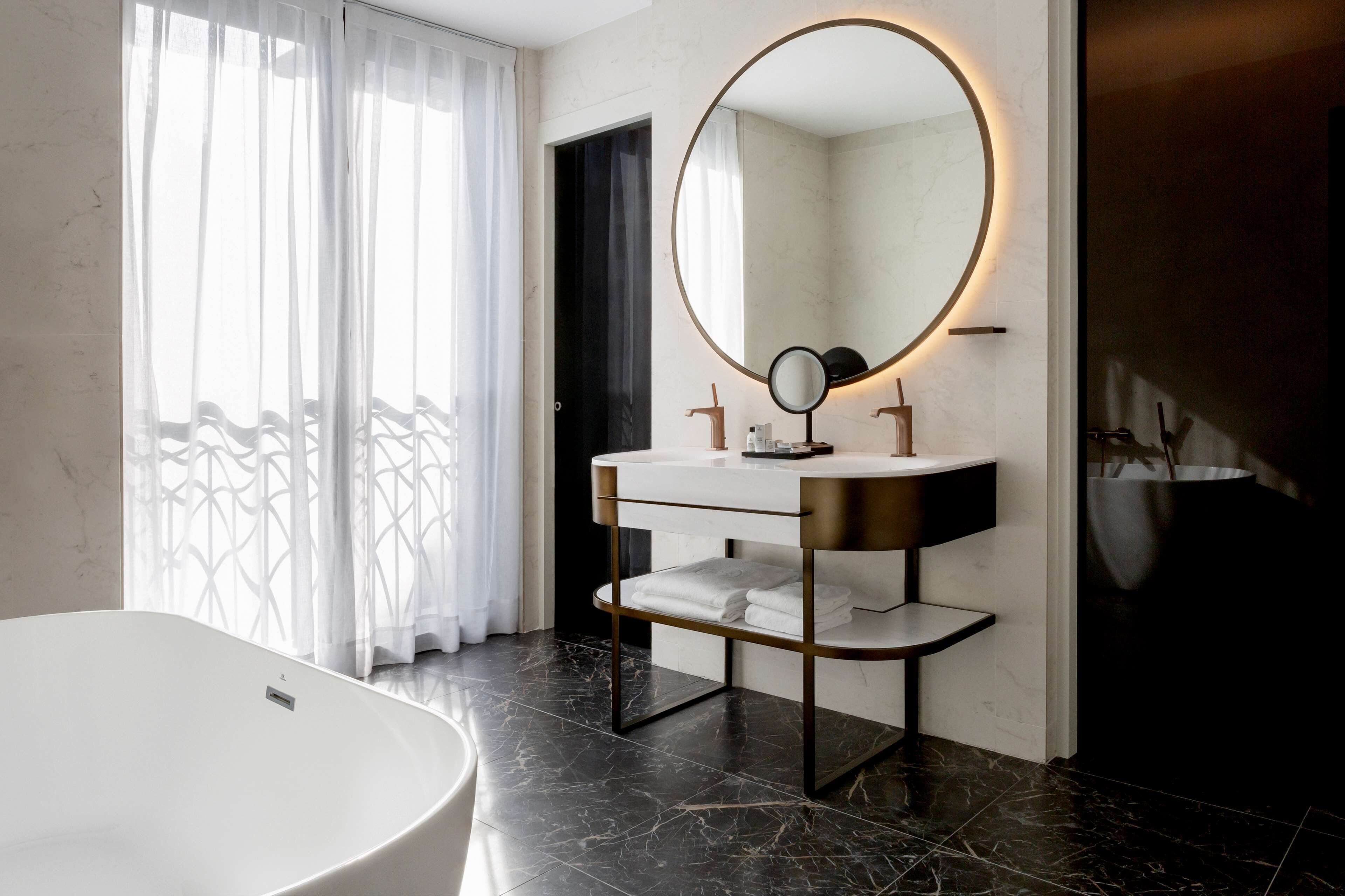 A modern bathroom with a freestanding bathtub, a vanity with a round mirror, and a dark tiled floor.