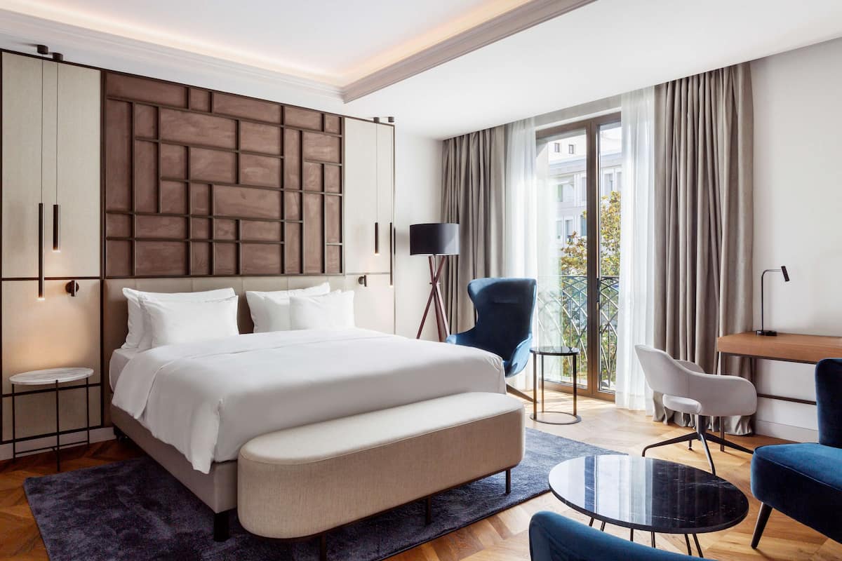A modern hotel room with a large bed, a desk, a chair, and a balcony with curtains.