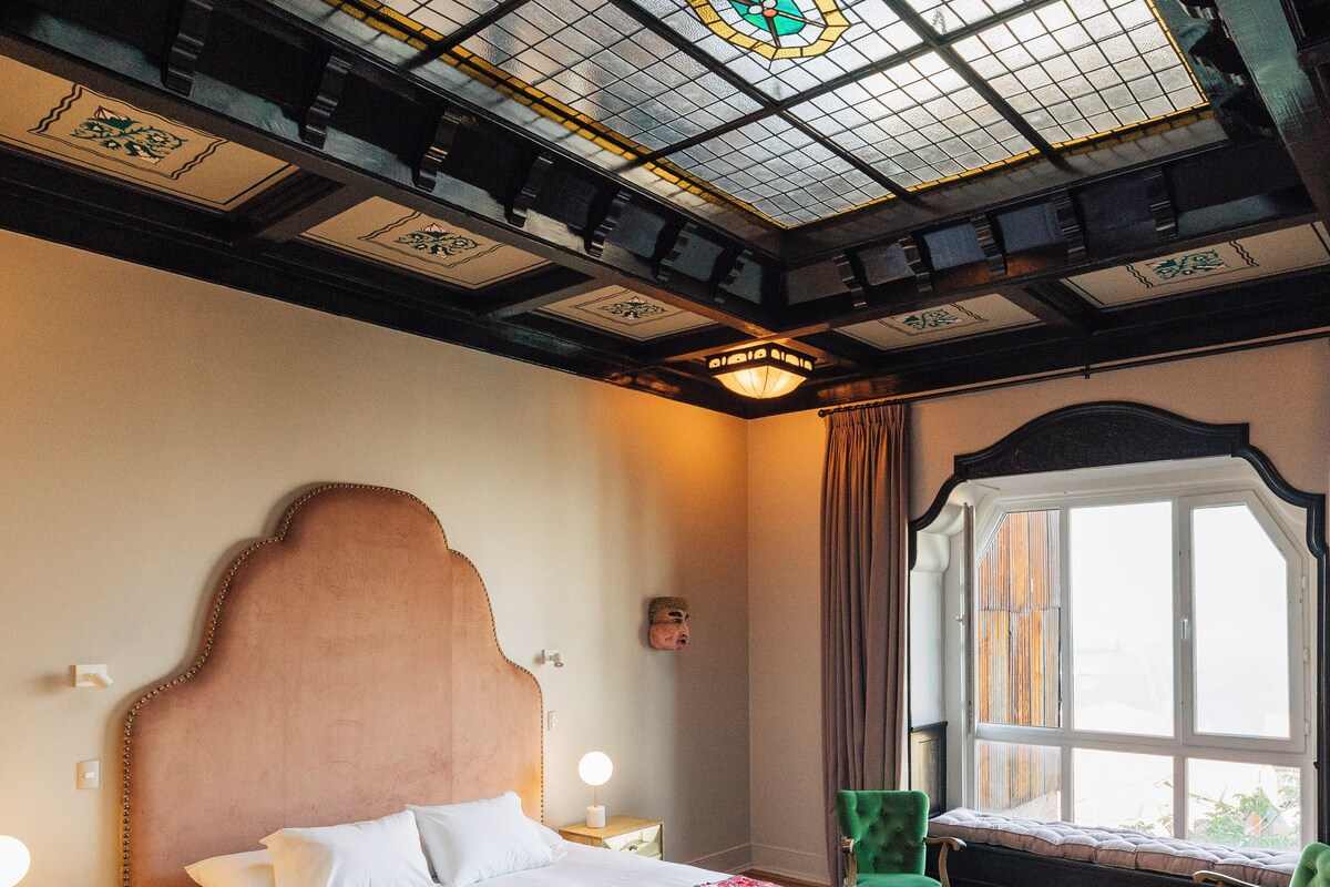 A hotel room with a large bed, a green armchair, a small table, and a large window.
