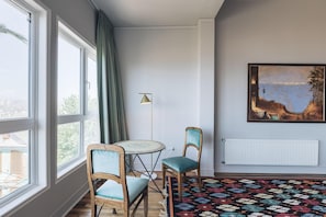 Habitació amb grans finestres, una taula rodona, dues cadires, una làmpada de peu, un radiador i un quadre a la paret.