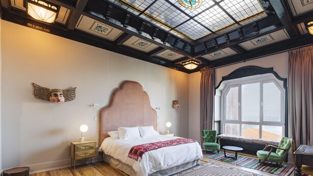 Un dormitorio con una cama grande, una mesita de noche, un sillón verde, una ventana con cortinas y un tragaluz.