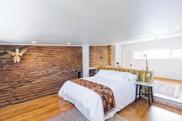 Une chambre avec un mur en briques, un plancher en bois, un lit recouvert d’une courtepointe blanche, un petit tabouret et une lampe.