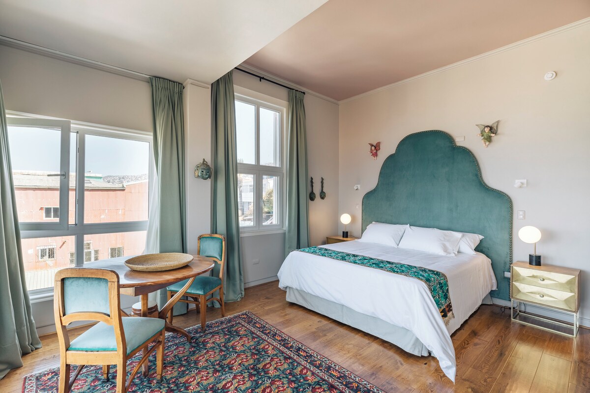 A bedroom with a bed, a table with chairs, a window with curtains, and a nightstand with a lamp.