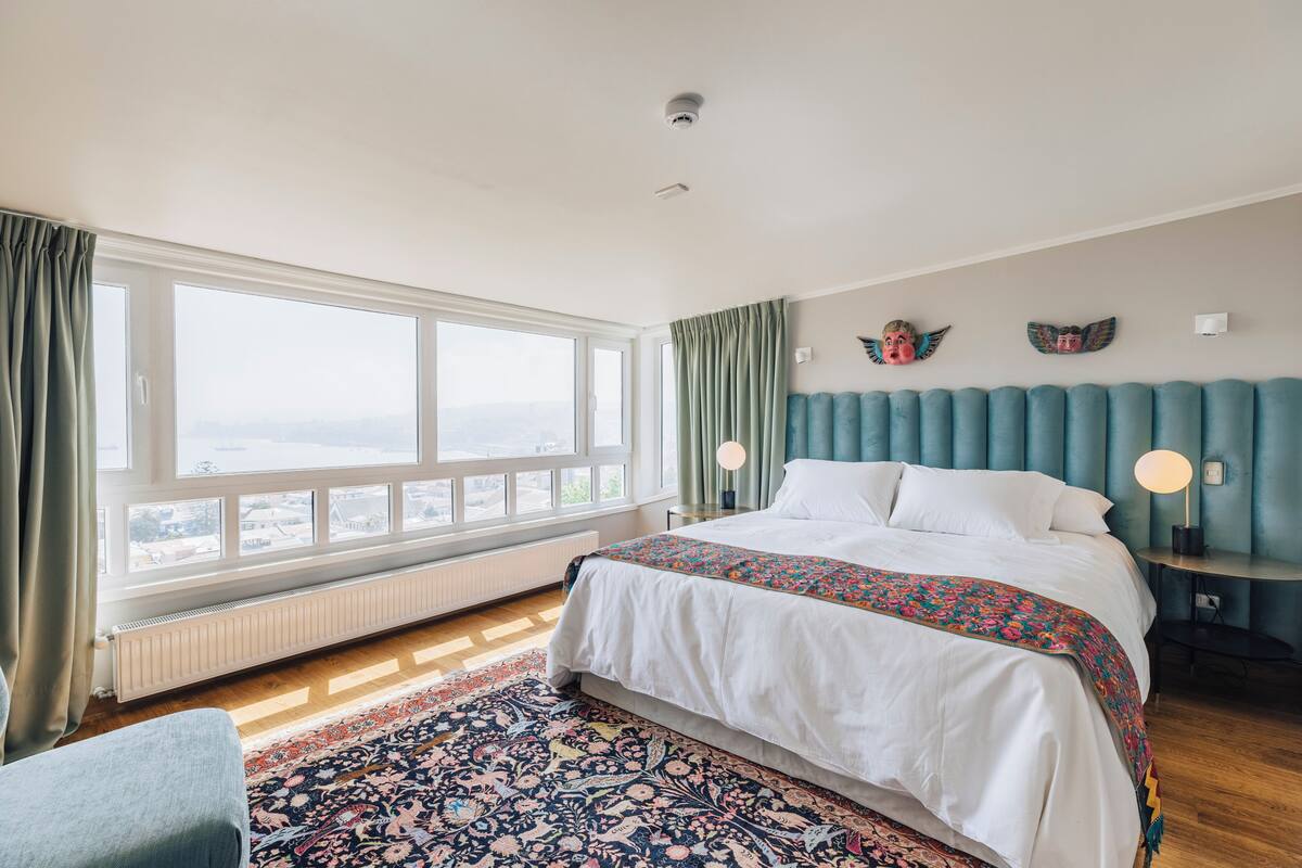 A bedroom with a large window, a bed with a floral bedspread, a blue upholstered headboard, and a nightstand with a lamp.