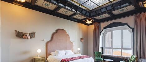Un dormitorio con una cama grande, una mesita de noche, un sillón verde, una ventana con cortinas y un tragaluz.