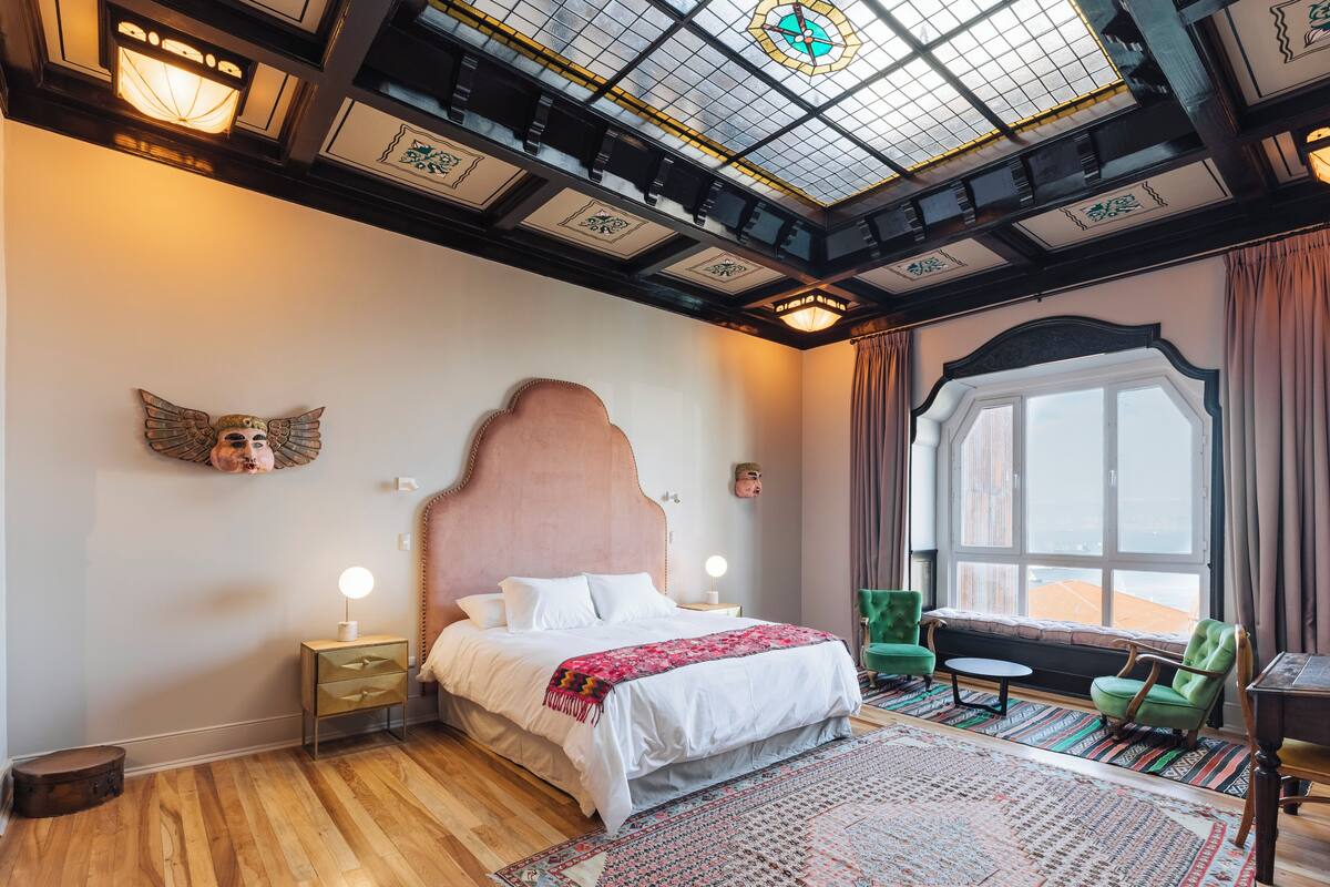 A bedroom with a large bed, a wooden floor, a skylight, a window with curtains, and a green chair.