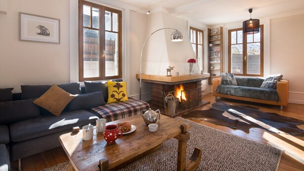 Una sala de estar acogedora con chimenea, un sofá, una mesa de centro con una taza y pasteles, y una estantería.
