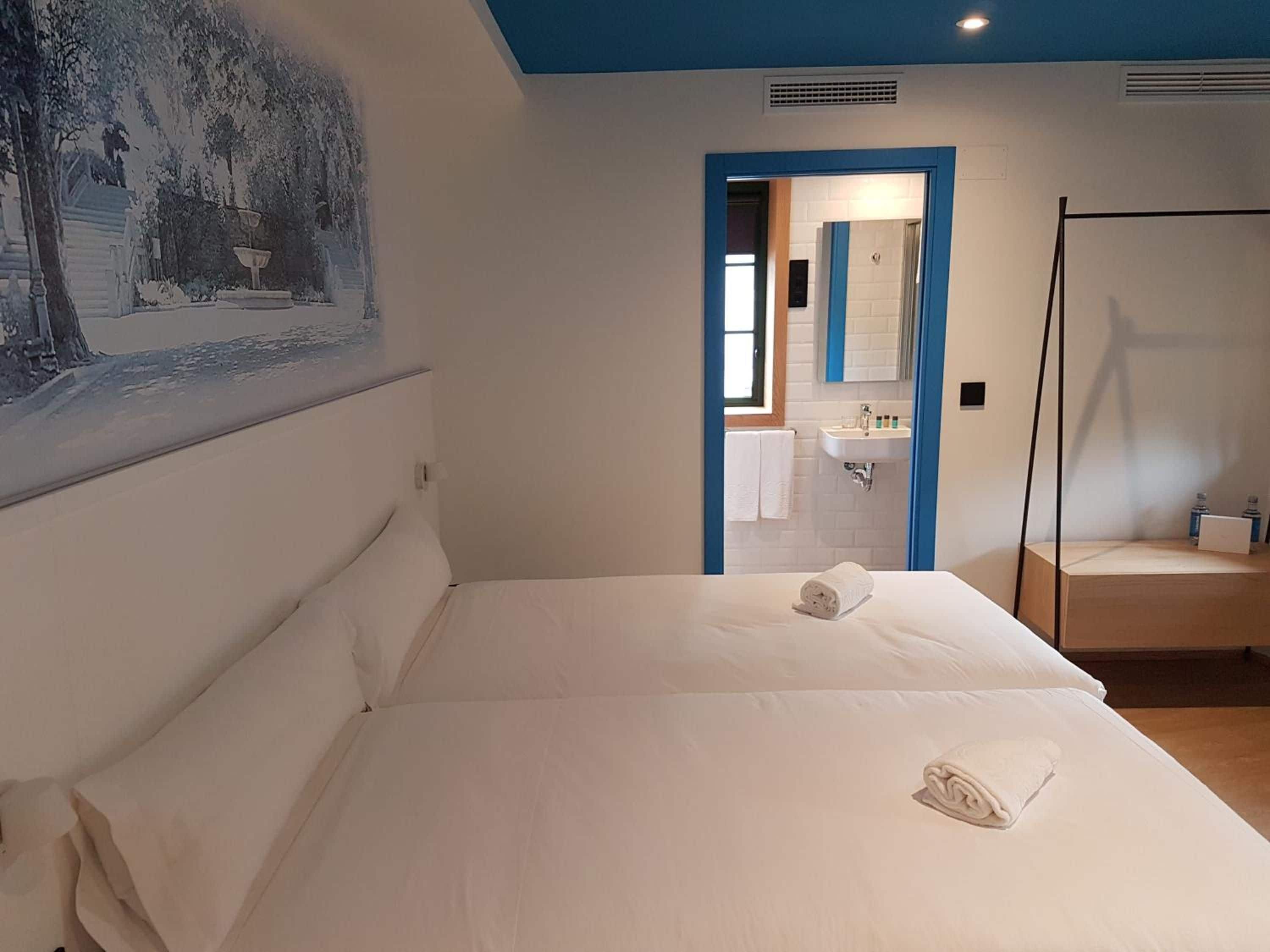 Een hotelkamer met een bed, een schilderij aan de muur, een badkamer zichtbaar door een open deur, en een nachtkastje met een lamp.