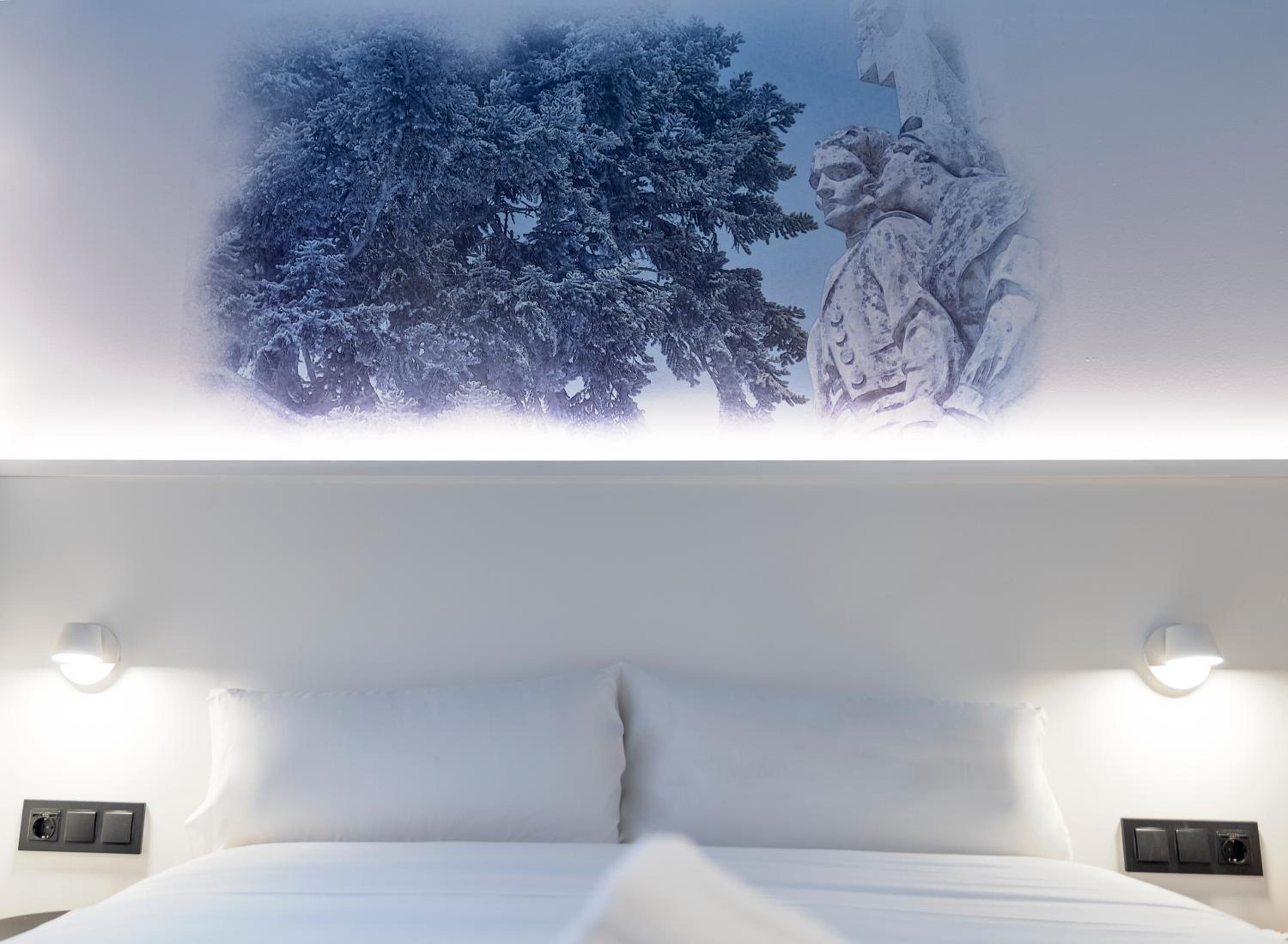A hotel room with a bed, two wall-mounted light fixtures, and a wall mural.