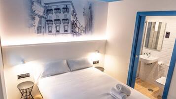 Una habitación de hotel con cama, mesitas de noche, un cuadro en la pared y un baño con lavamanos y espejo.