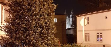 A nighttime street scene with a building featuring a large tree in the foreground and a church with illuminated spires in the background.