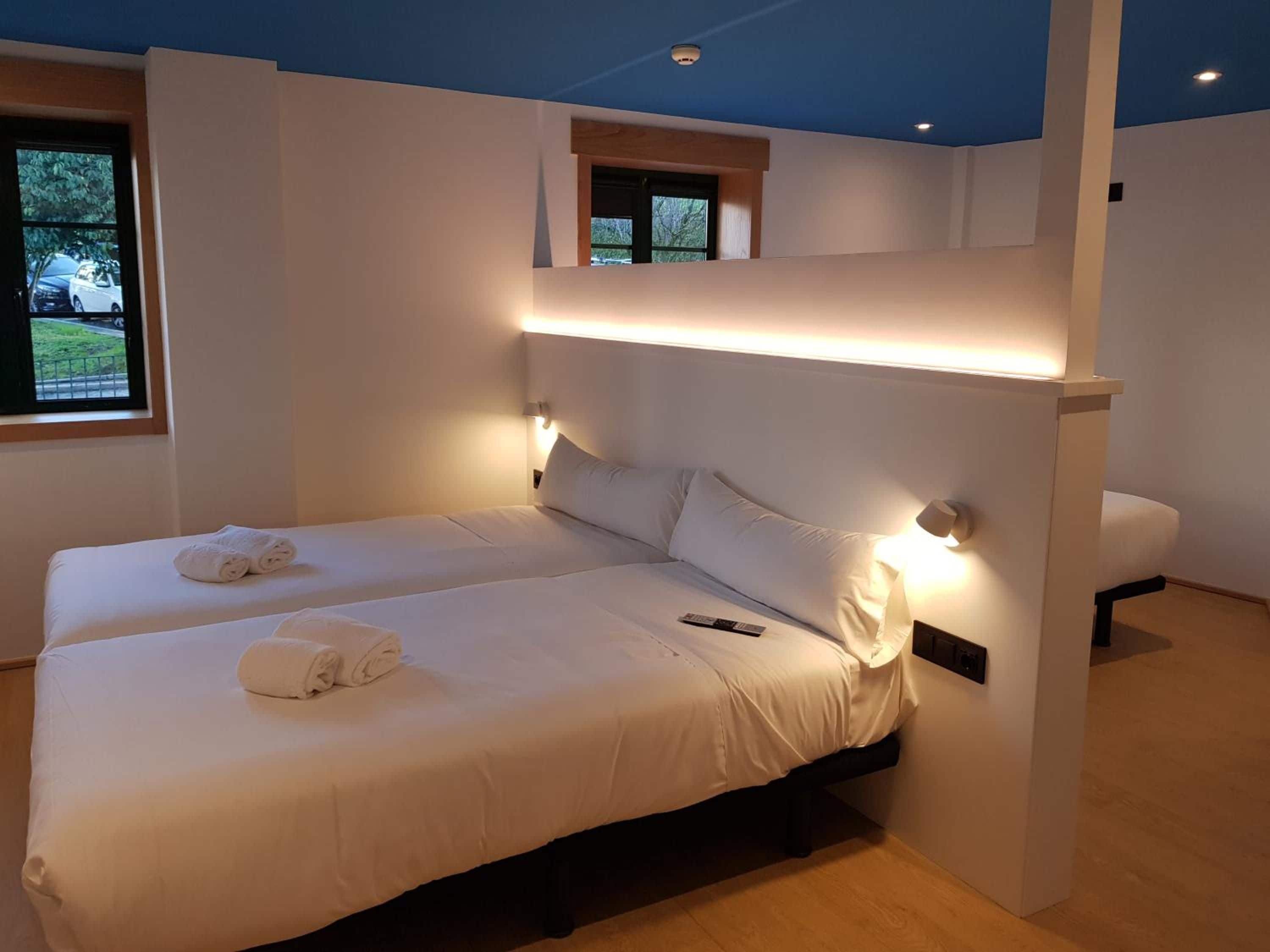A modern hotel room with two beds, a small window, and a view of the outdoors.