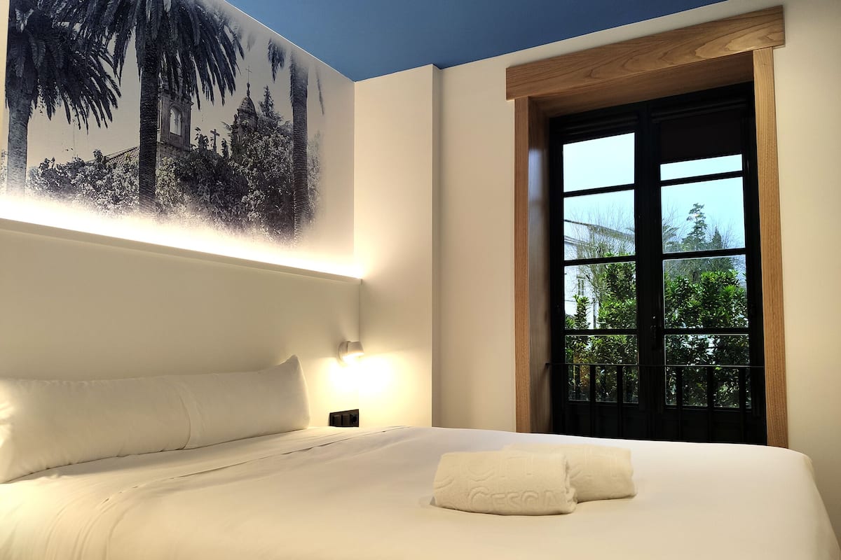 Habitación de hotel con una cama grande, una mesita de noche, una lámpara de pared y una ventana con vistas a zonas verdes.