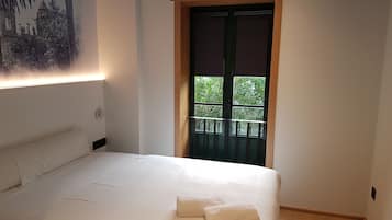 A bedroom with a bed, two towels, a nightstand, a wall-mounted lamp, a window with a view of greenery, and a wooden ceiling trim.