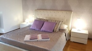 Ein ordentlich bezogenes Bett mit einem gemusterten Überwurf und zwei violetten Kissen, flankiert von weißen Nachttischen mit Lampen.