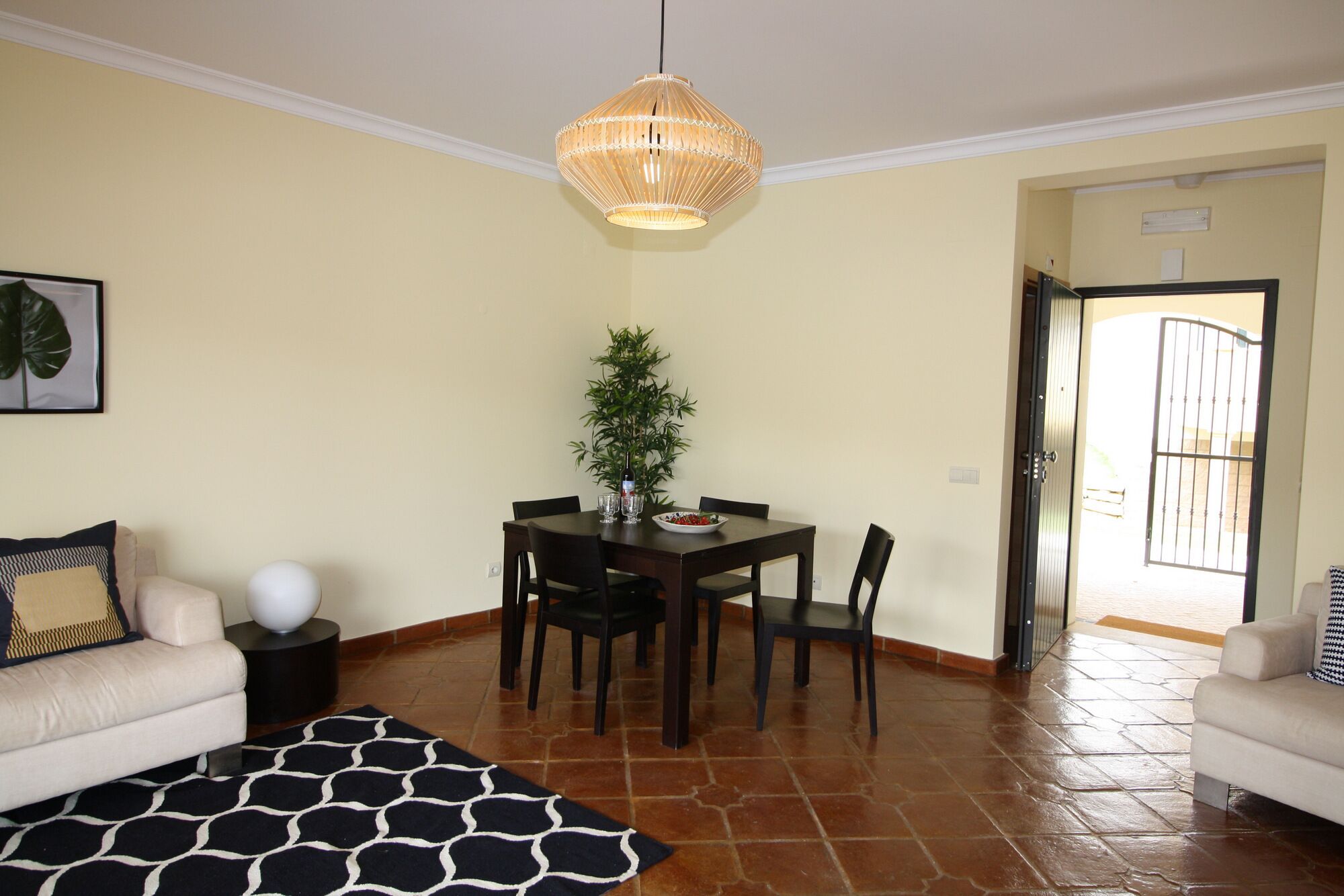 A dining area with a table and chairs, a hanging light fixture, a potted plant, and a sofa with a patterned rug.