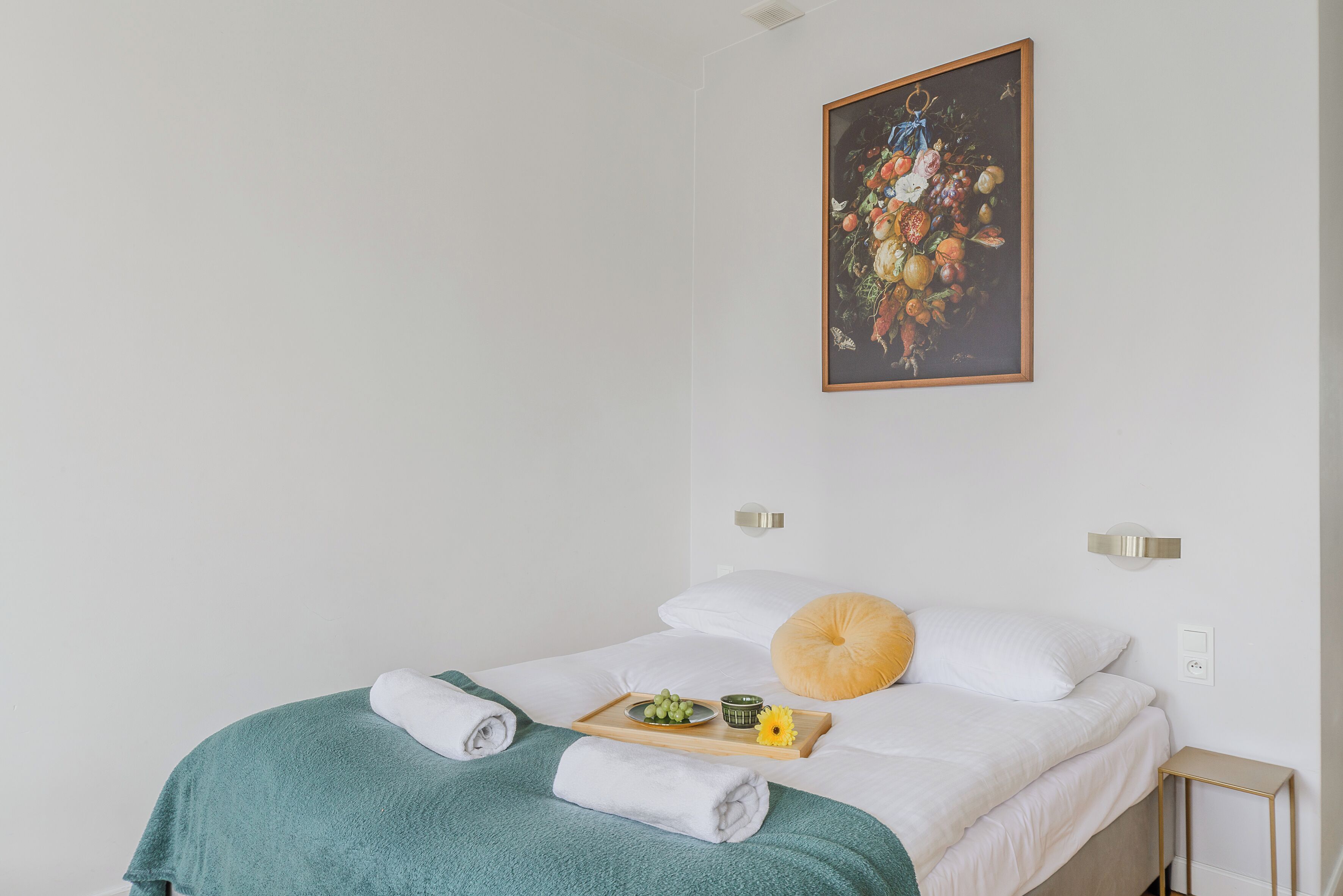 A neatly made bed with a green towel, a tray with fruits and a small bowl, and a painting on the wall.