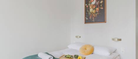 A neatly made bed with a green towel, a tray with fruits and a small bowl, and a painting on the wall.