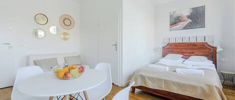 Un dormitorio con cama de madera, sillas blancas, una mesa redonda con fruta y decoración en la pared.